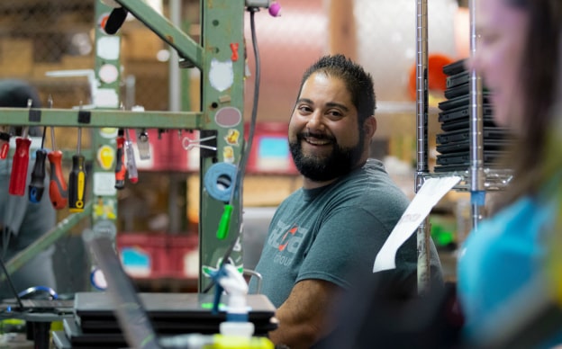 ARCOA employee smiling at coworker in workshop with various tools and electronics